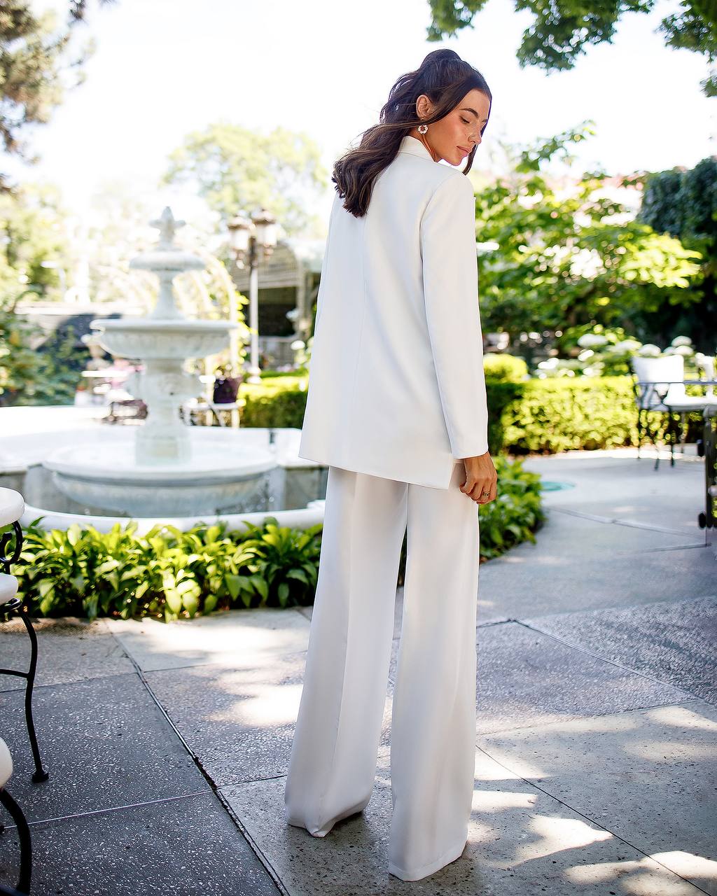Woman in a white outfit standing in an outdoor setting with greenery and a fountain.