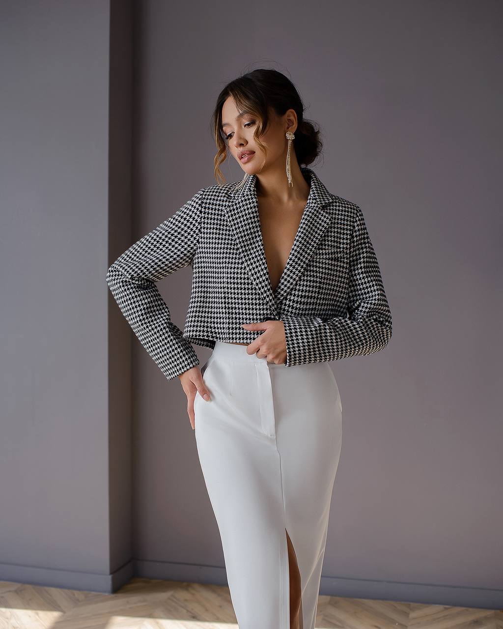 Woman wearing a black and white checkered blazer and white pants against a gray wall.