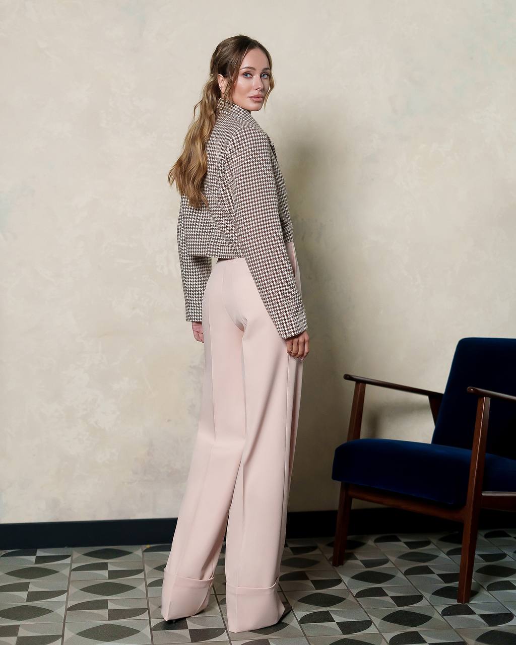 Woman in a checkered shirt and light pink pants standing in a room with a blue chair and patterned floor.