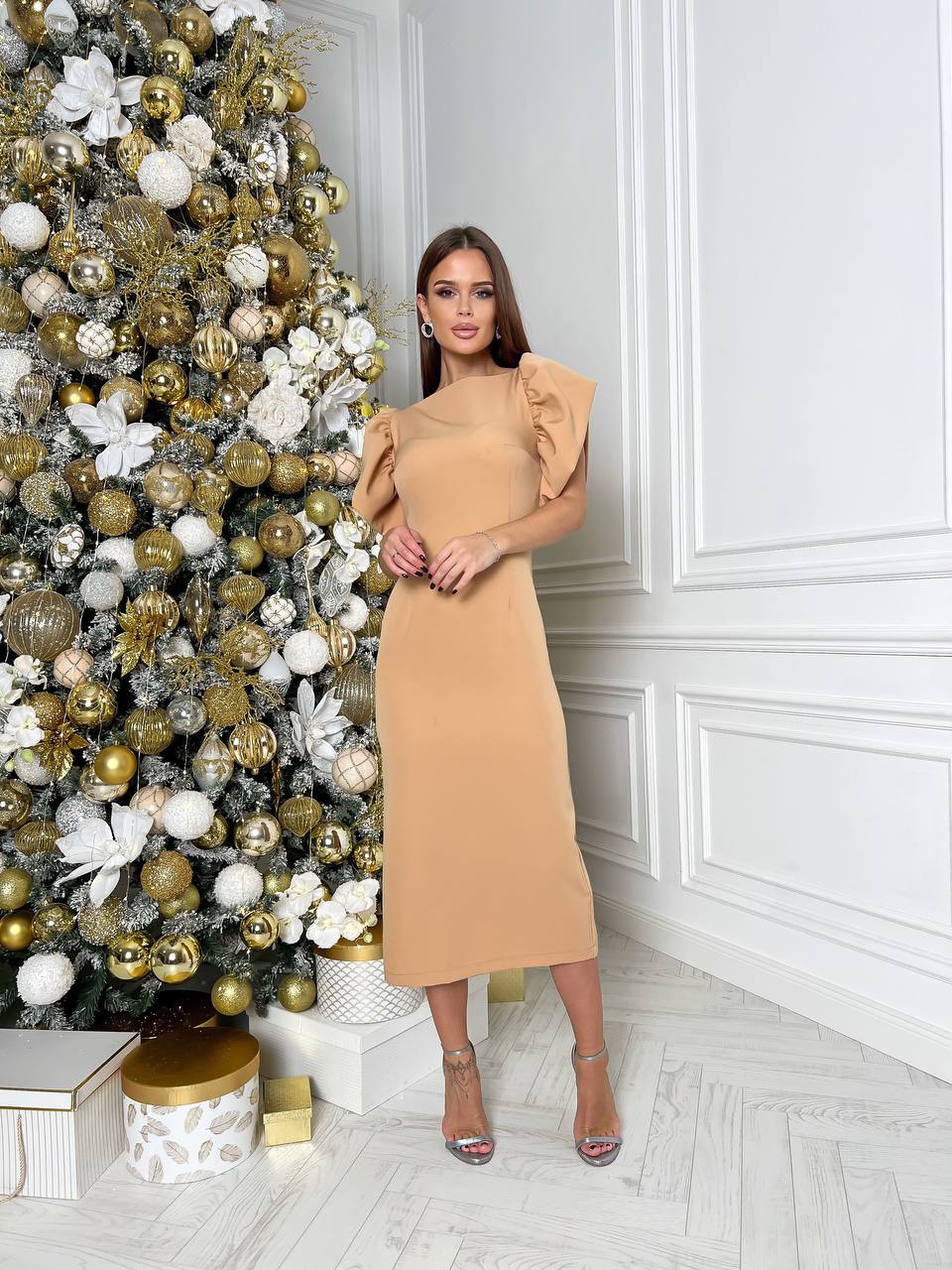 Woman in a beige dress standing next to a decorated Christmas tree in a room with white walls.