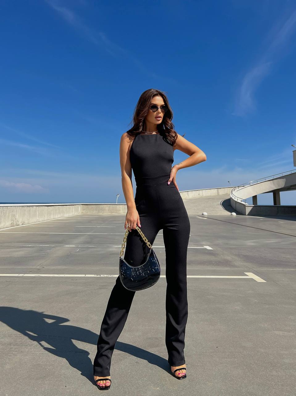 Woman in a black outfit holding a blue handbag on a rooftop with a clear blue sky.