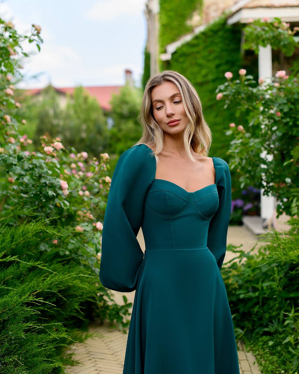 Woman in a teal dress standing in a garden with flowers and greenery.