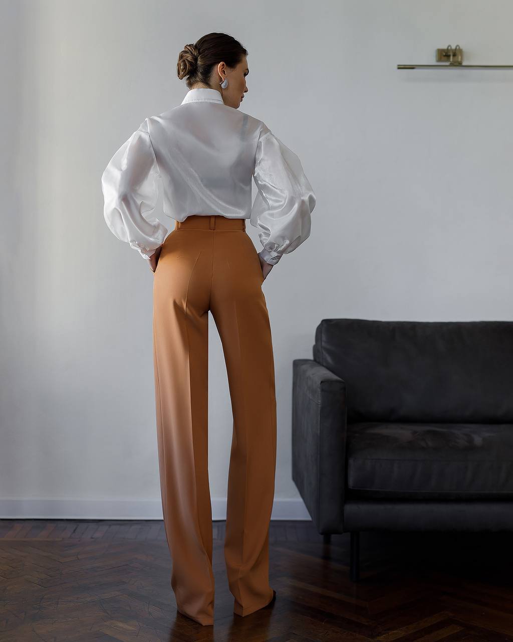 Person wearing a white blouse and brown pants standing in a room with a gray sofa.