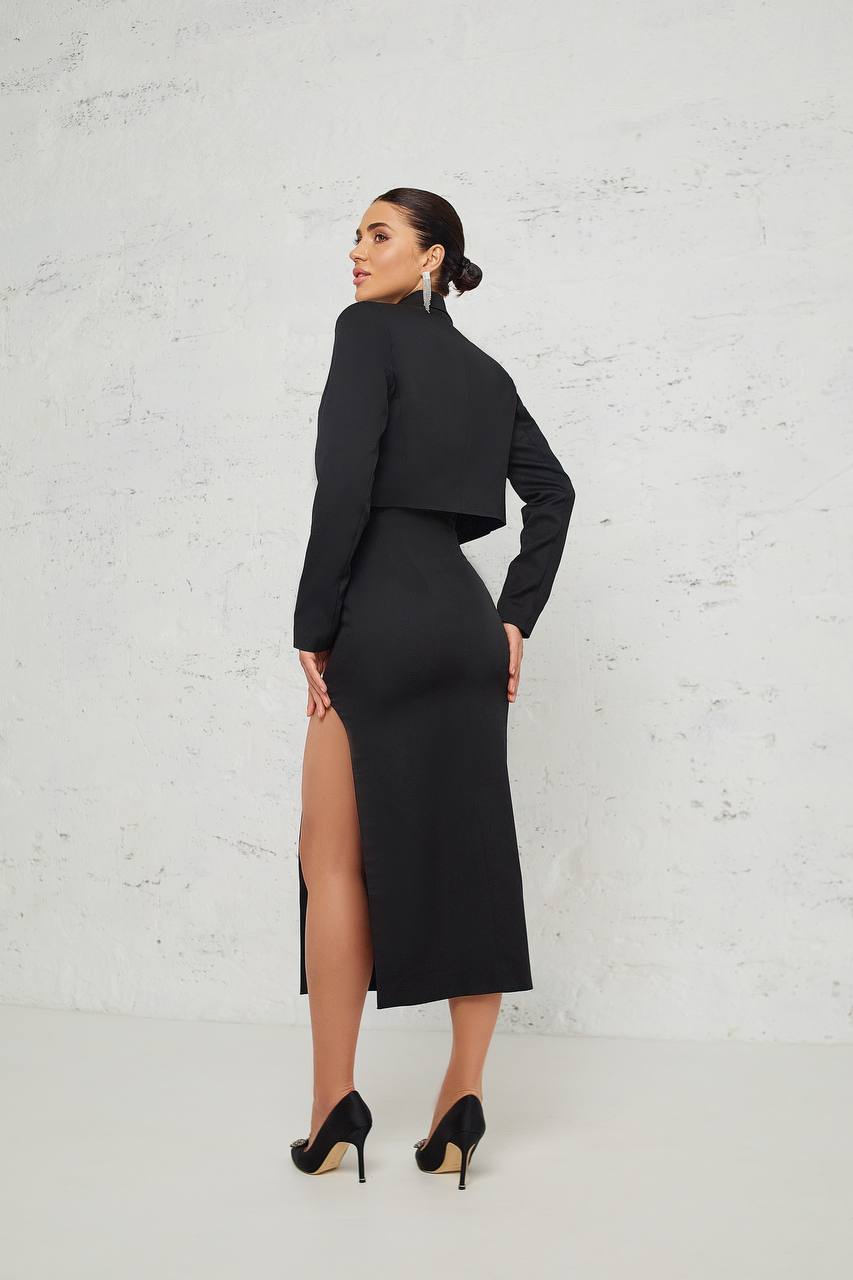 Woman wearing a black outfit with a high slit skirt against a white brick wall.