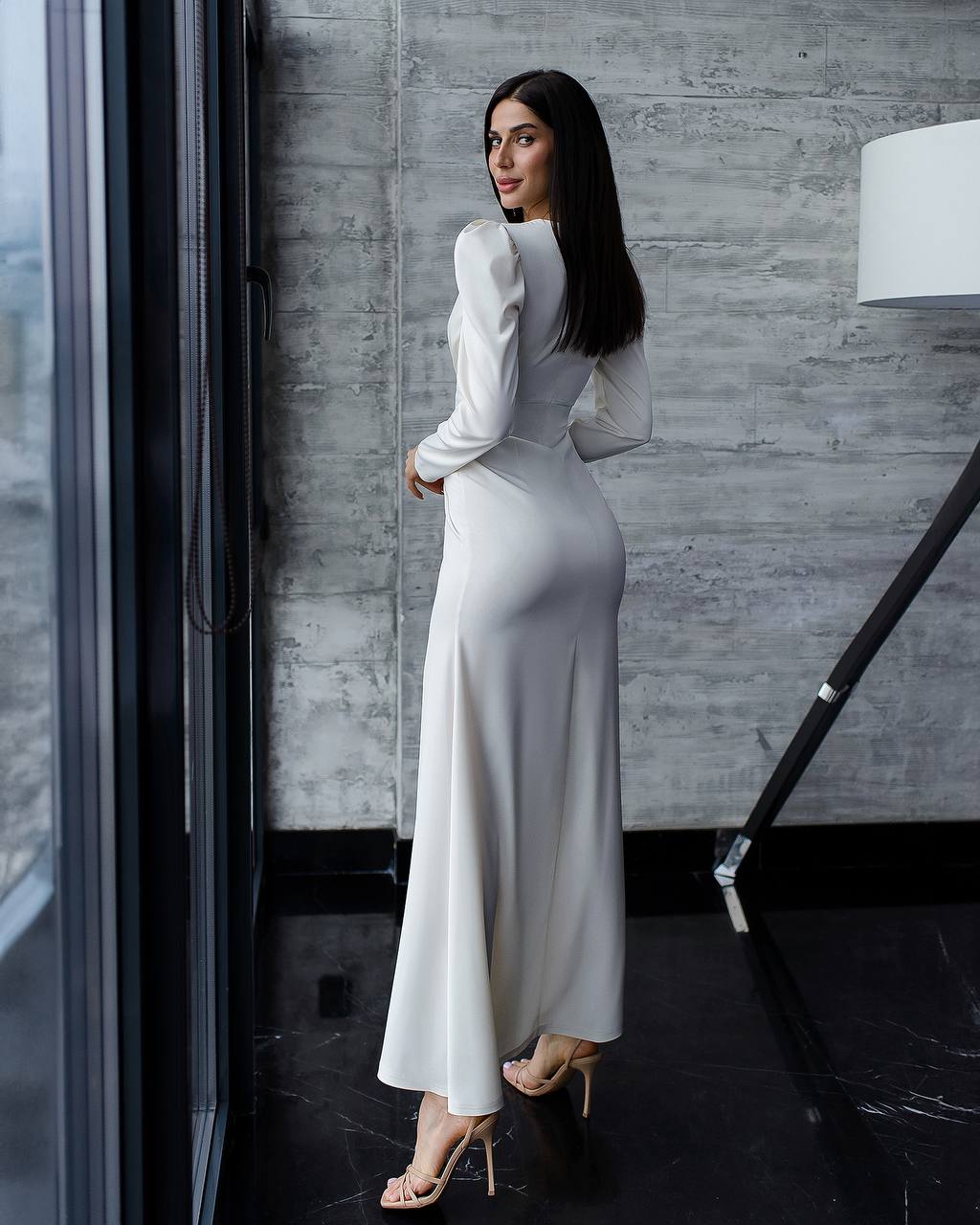 Woman in a long, light-colored dress standing indoors against a gray wall.