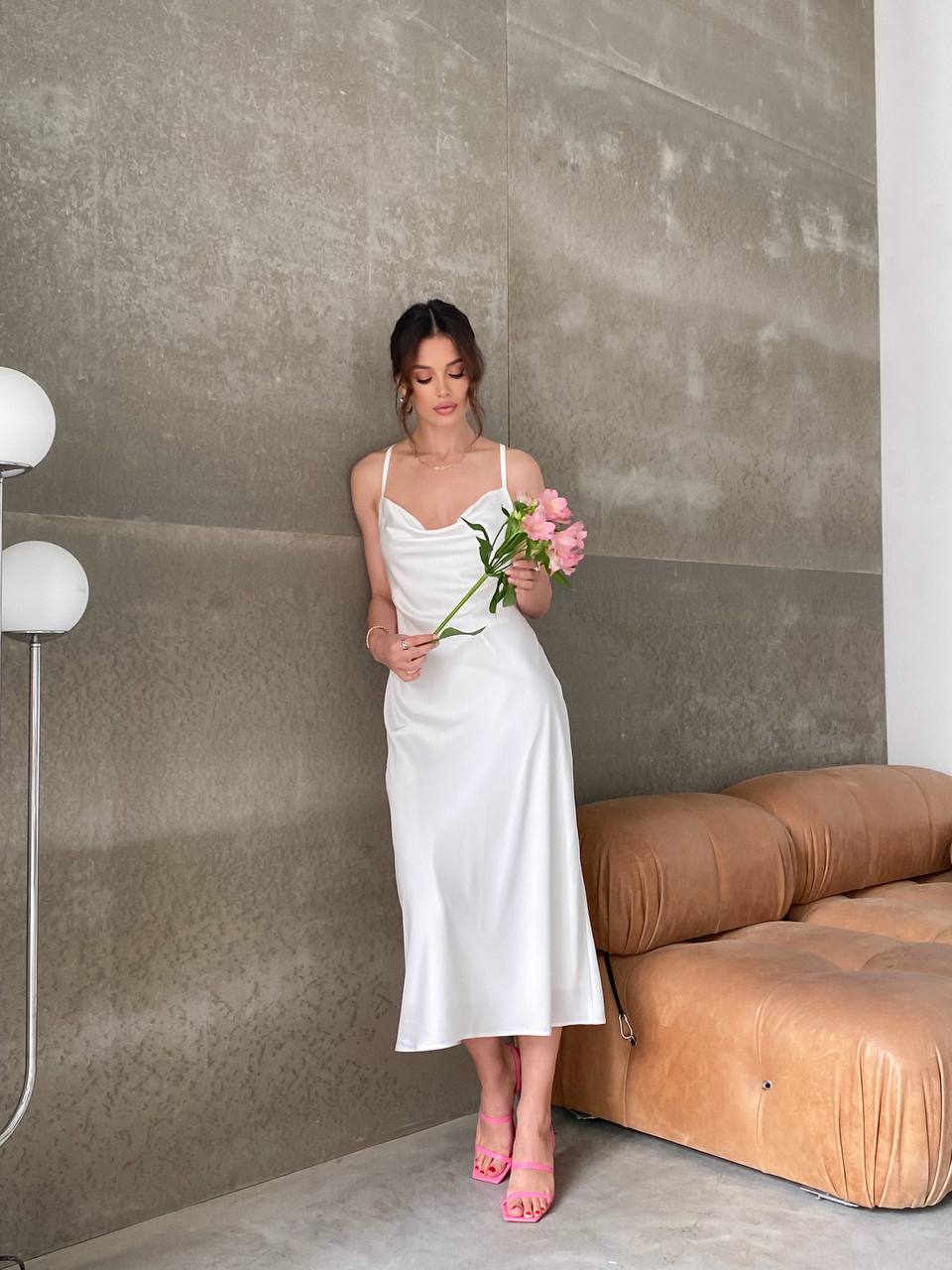 Woman in a white dress holding flowers in a modern room with a brown sofa and concrete wall.