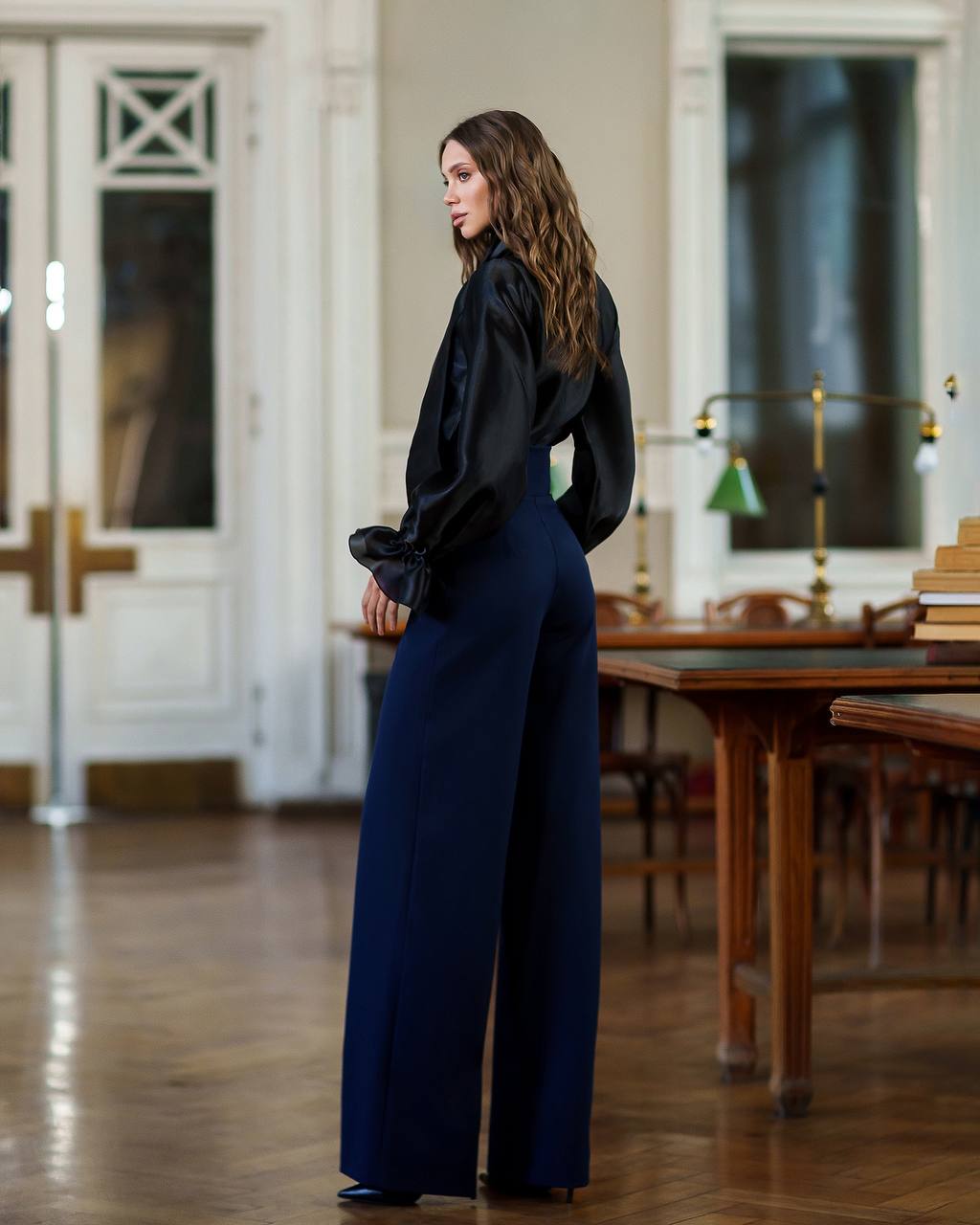 Woman in a black top and blue pants standing in an elegant room with wooden tables and chairs.