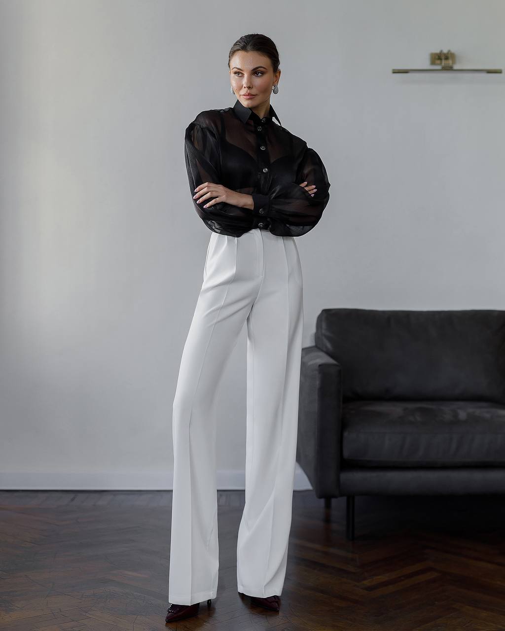 Woman in black sheer blouse and white pants standing in a room with a gray sofa.