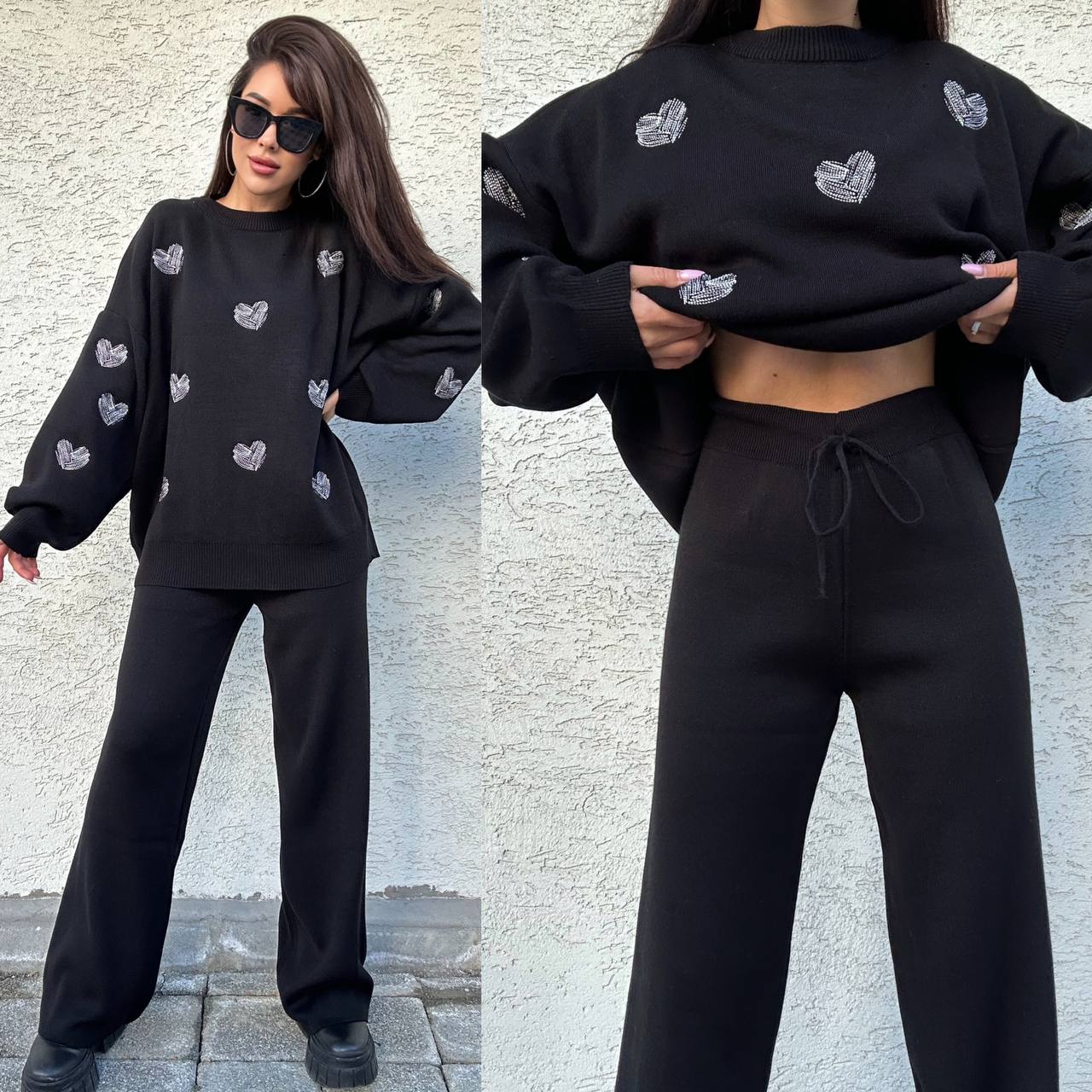 Woman wearing a black outfit with heart patterns, standing against a textured wall.