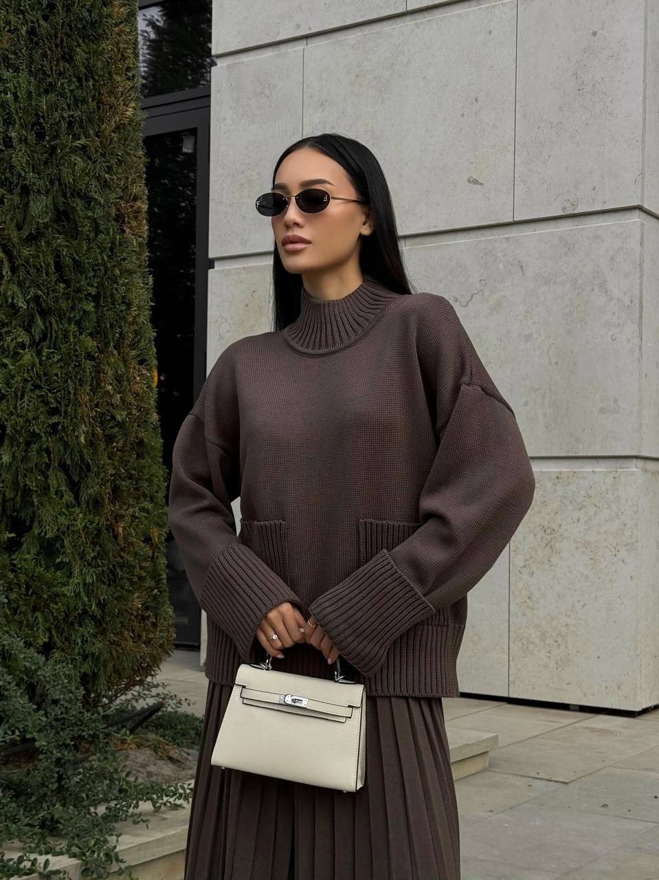 Woman wearing a brown sweater and skirt with a white handbag, standing against a stone wall.