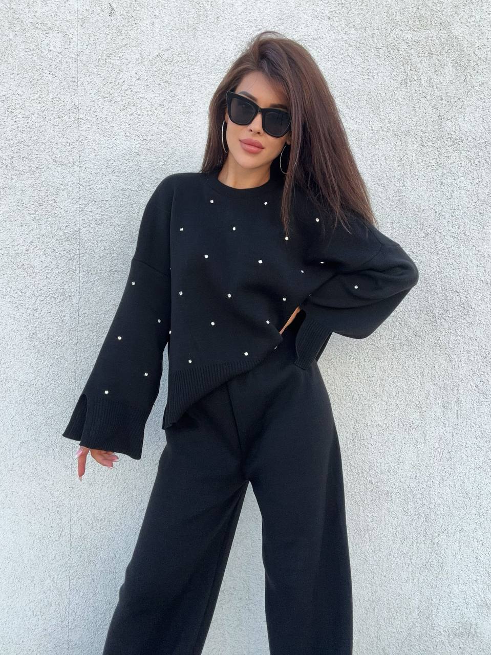 Woman wearing a black outfit with small embellishments against a light gray wall.