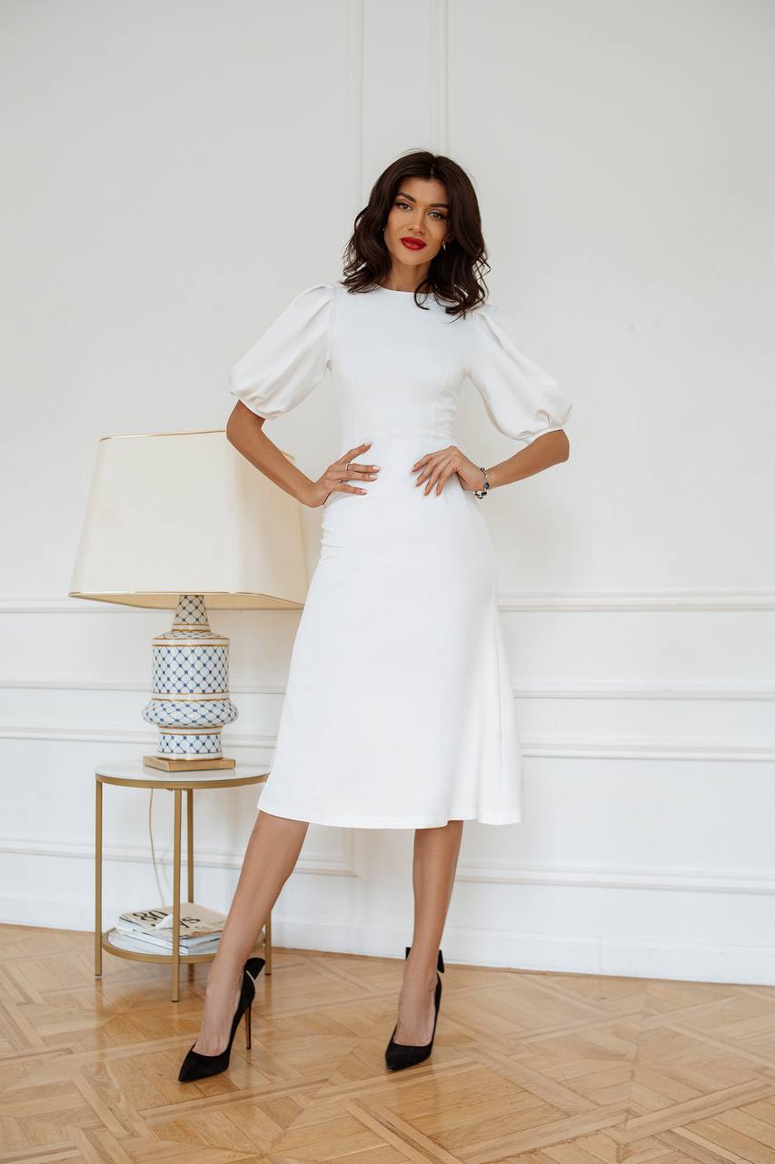Woman in a white dress standing in a room with a lamp and decorative items.