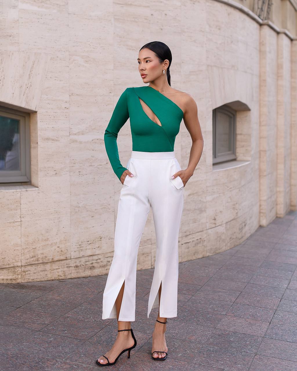 Woman wearing a green top and white pants standing against a beige wall.