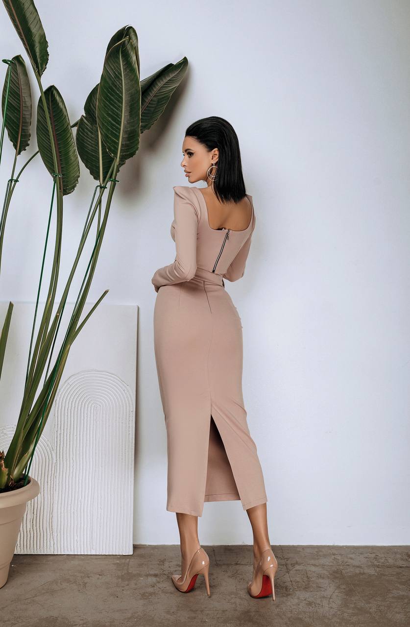 Woman in a beige dress standing next to a large plant indoors.