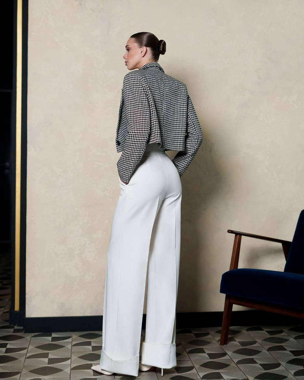 Woman wearing a black and white checkered top and white pants standing in a room with a chair and patterned floor.
