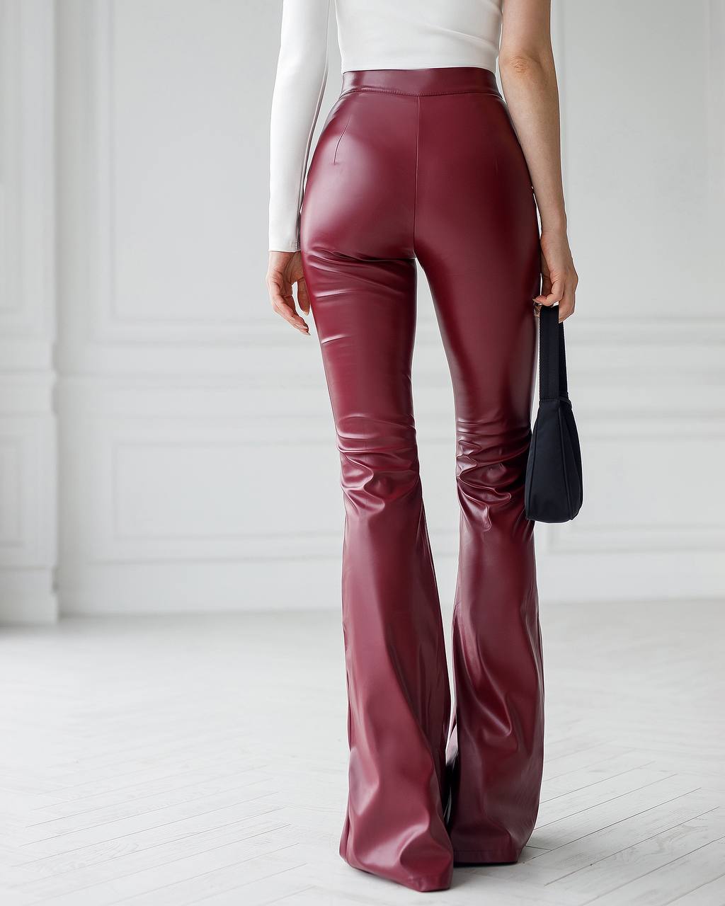 Person wearing maroon vinyl pants with a white top and black handbag on a light wooden floor.