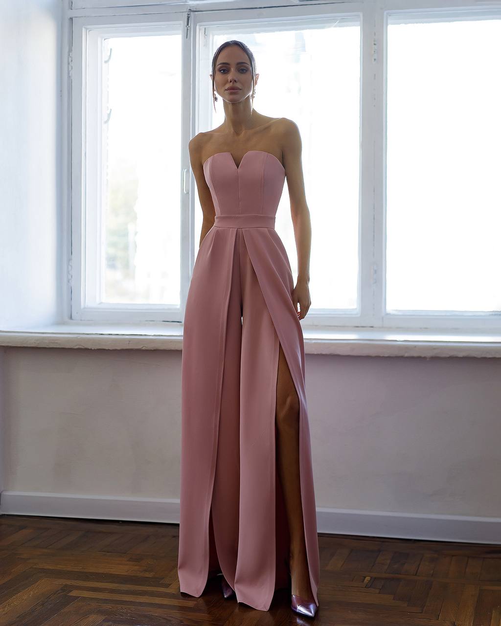 Woman wearing a pink strapless gown with a slit standing in front of a window.