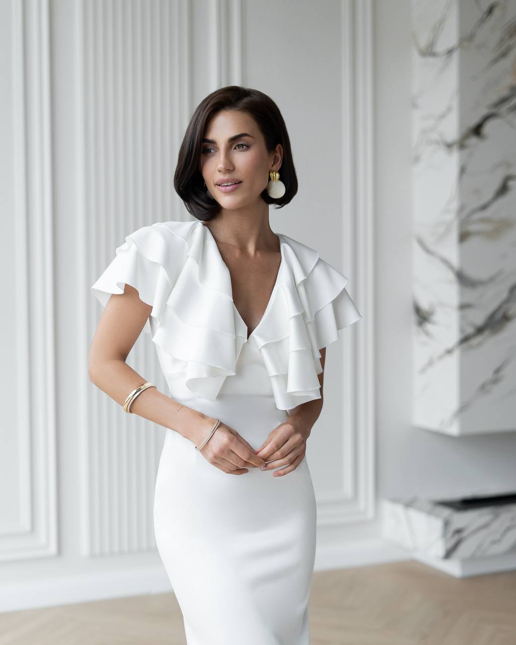 Woman wearing a white dress with ruffled details in a room with marble walls.
