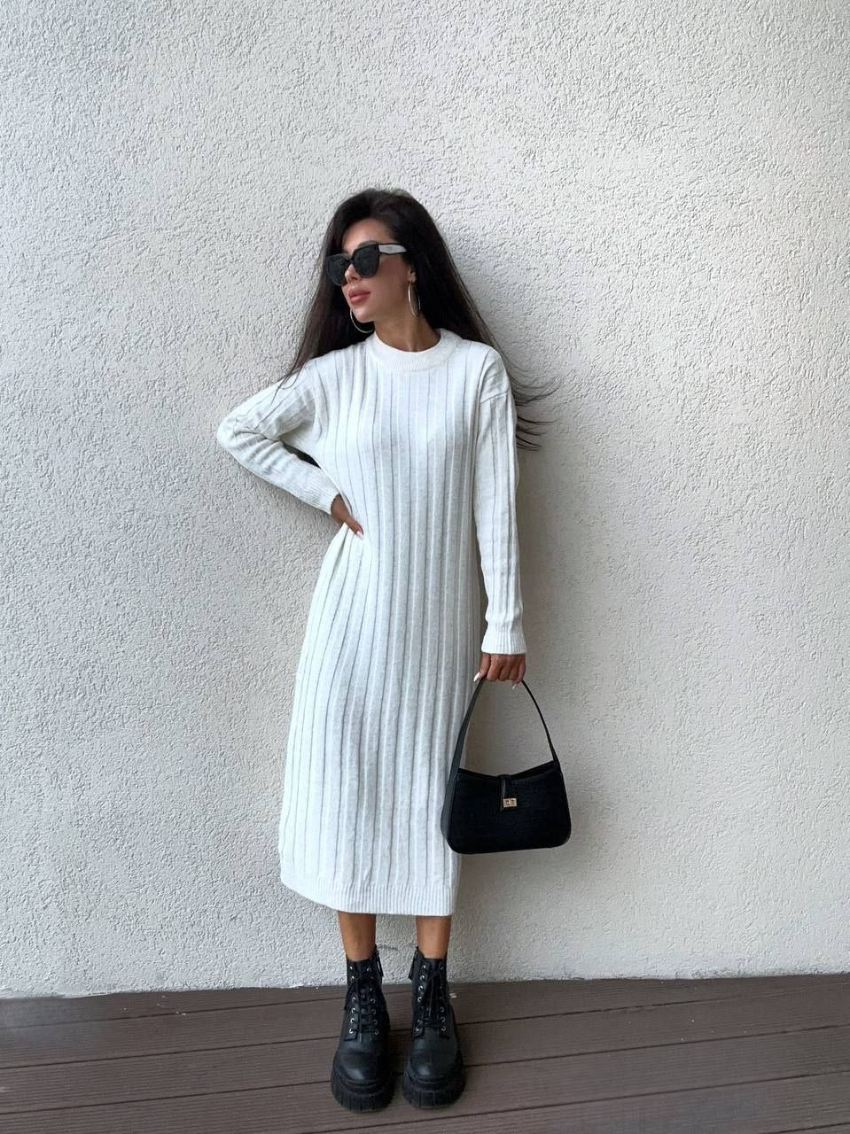 Woman wearing a white ribbed knit dress with black boots and a black handbag against a plain wall.