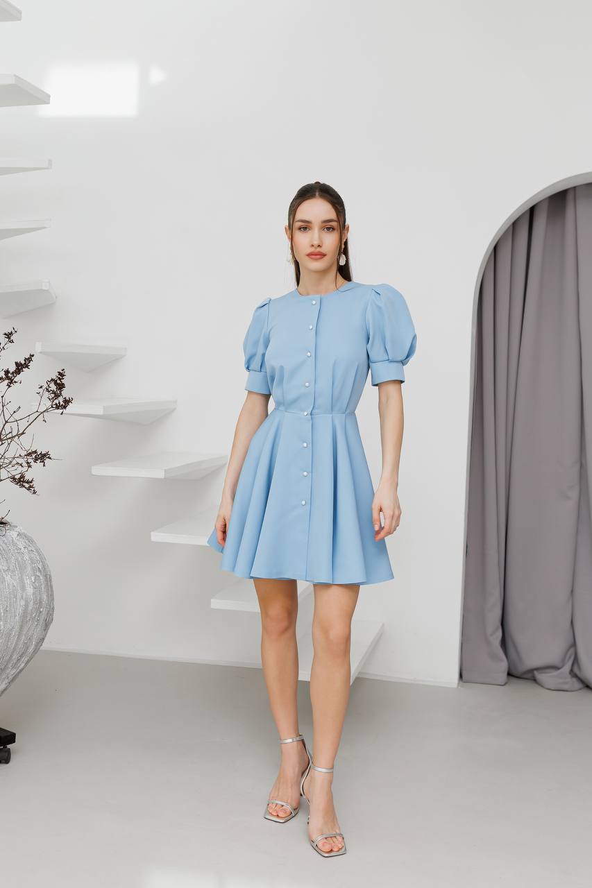 Woman wearing a light blue dress standing in a minimalistic room with white walls and gray curtains.