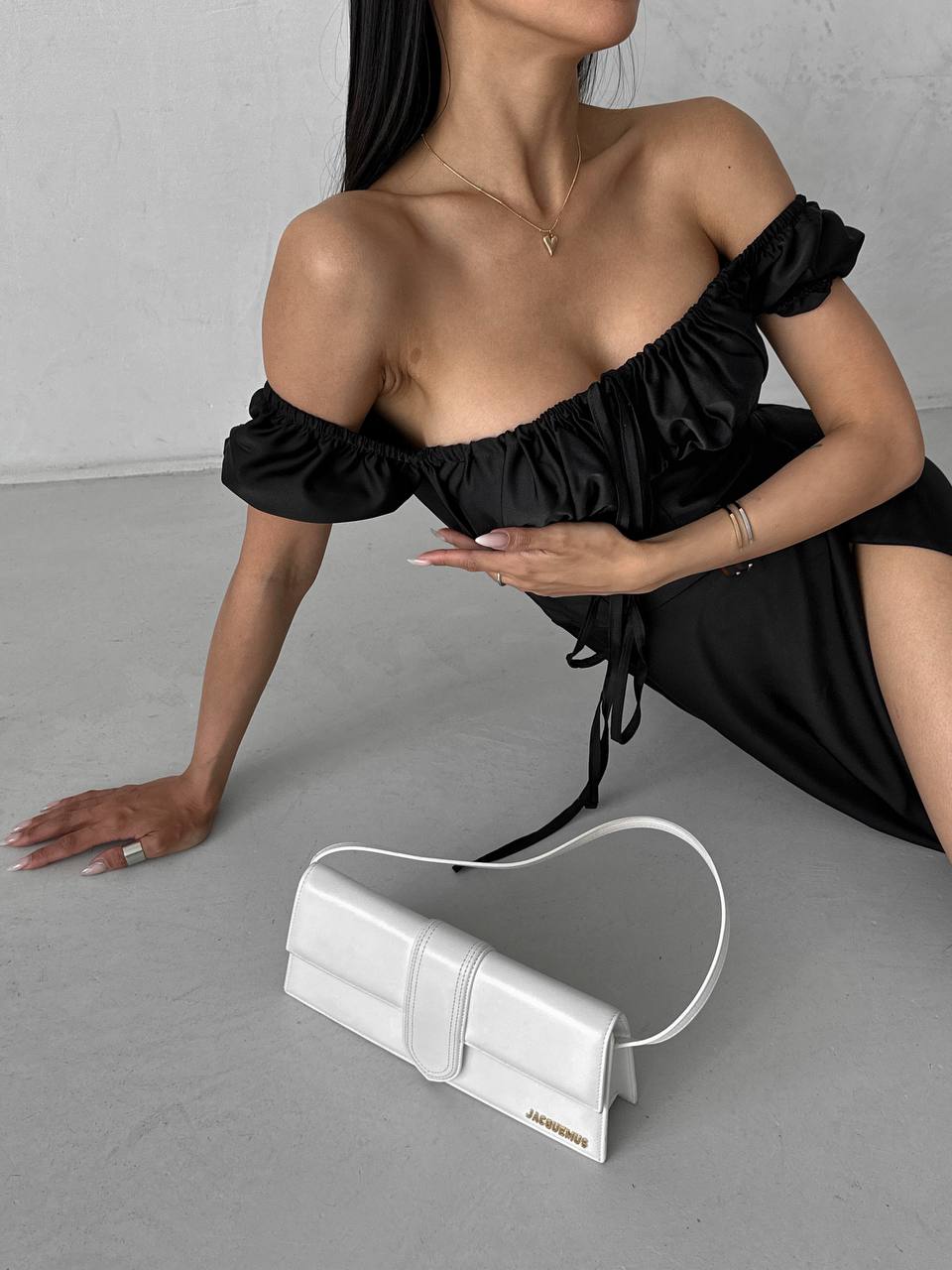 Woman in black off-shoulder dress with a white handbag on a gray floor.