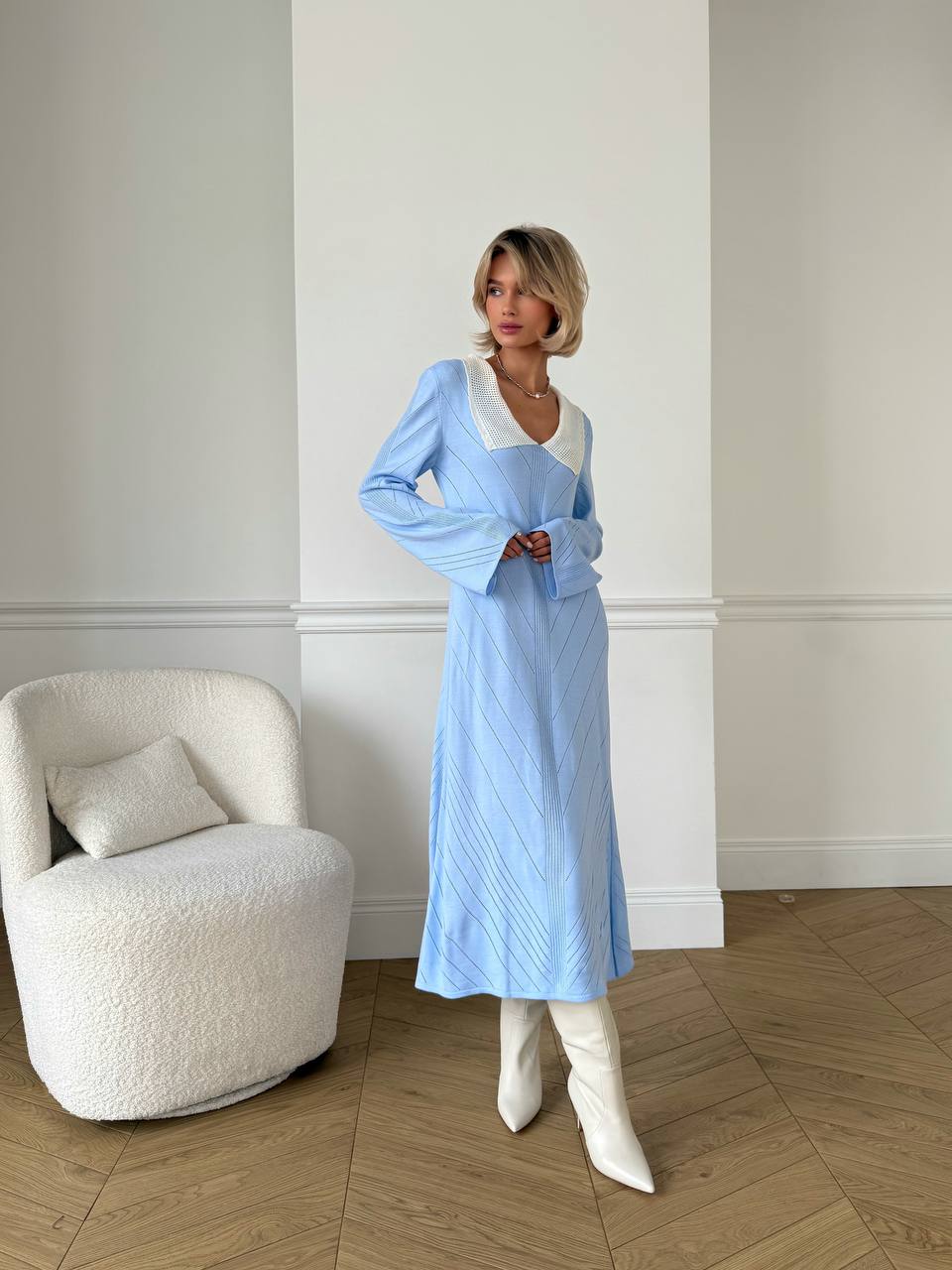 Woman in a light blue dress standing next to a white armchair in a room with wooden flooring.