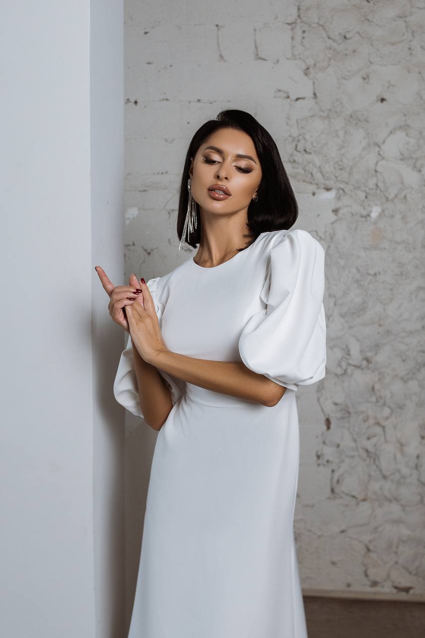Woman in a white dress with puffed sleeves against a textured wall.