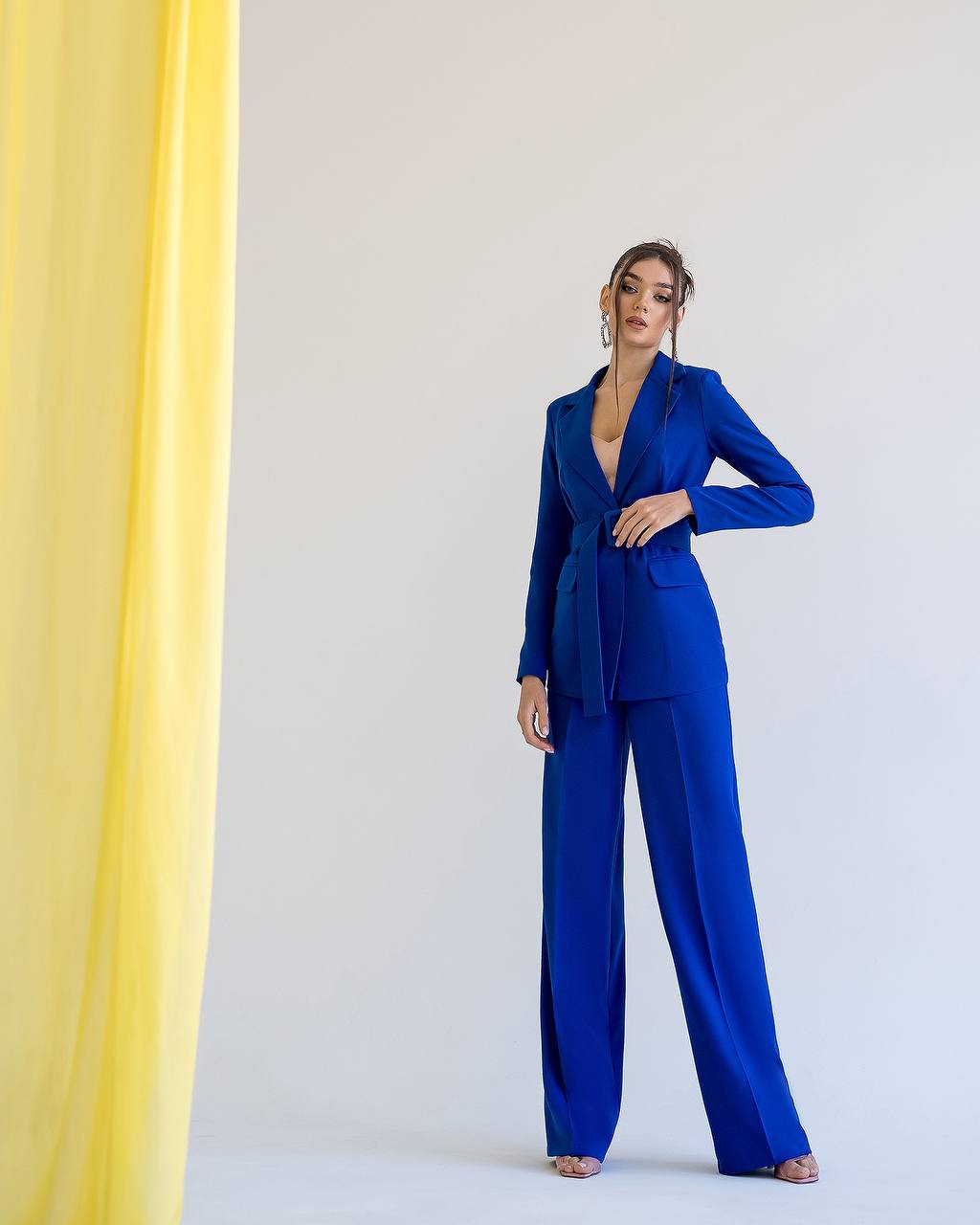 Woman wearing a blue suit standing against a white background with a yellow curtain on the side.