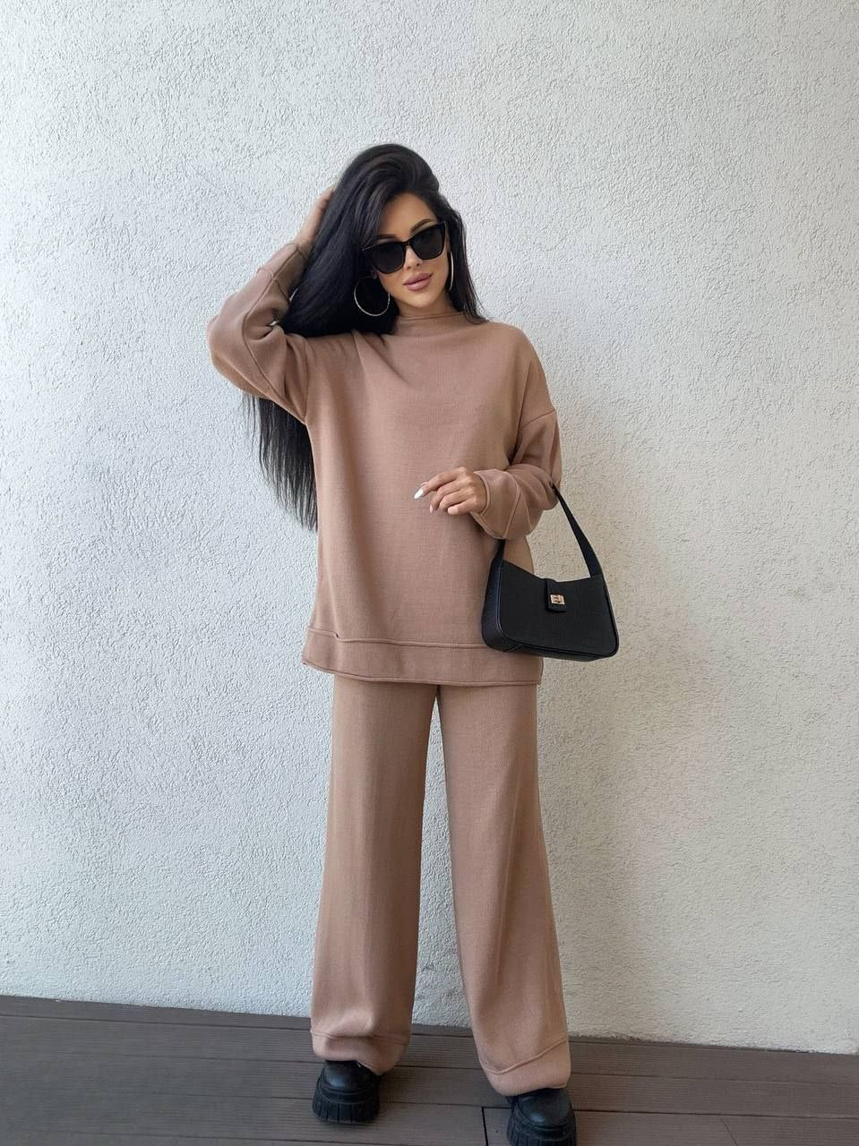 Woman wearing a beige outfit with sunglasses and a black handbag against a white wall.