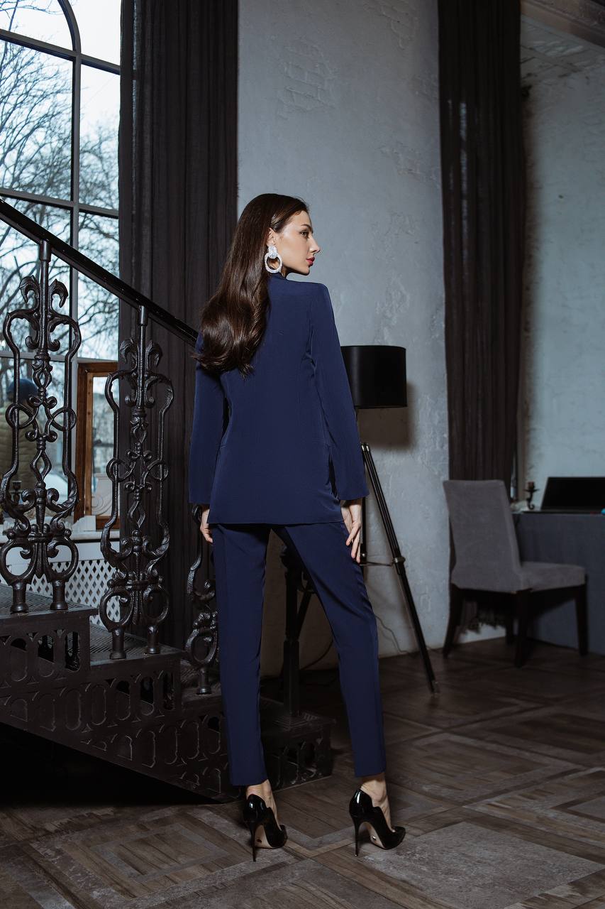 Woman in a navy blue suit standing in an elegant room with large windows and decorative elements.