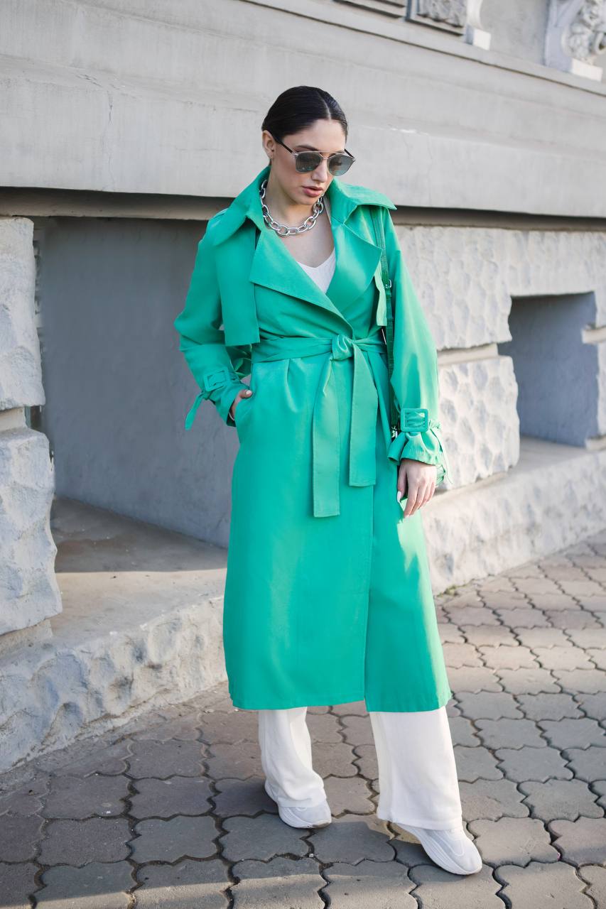 Woman wearing a green trench coat and white outfit standing on a street.