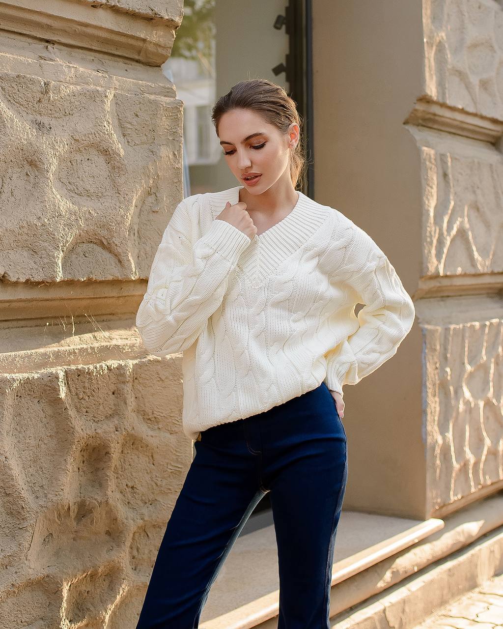 Woman wearing a white cable knit sweater and dark blue pants standing against a stone wall.