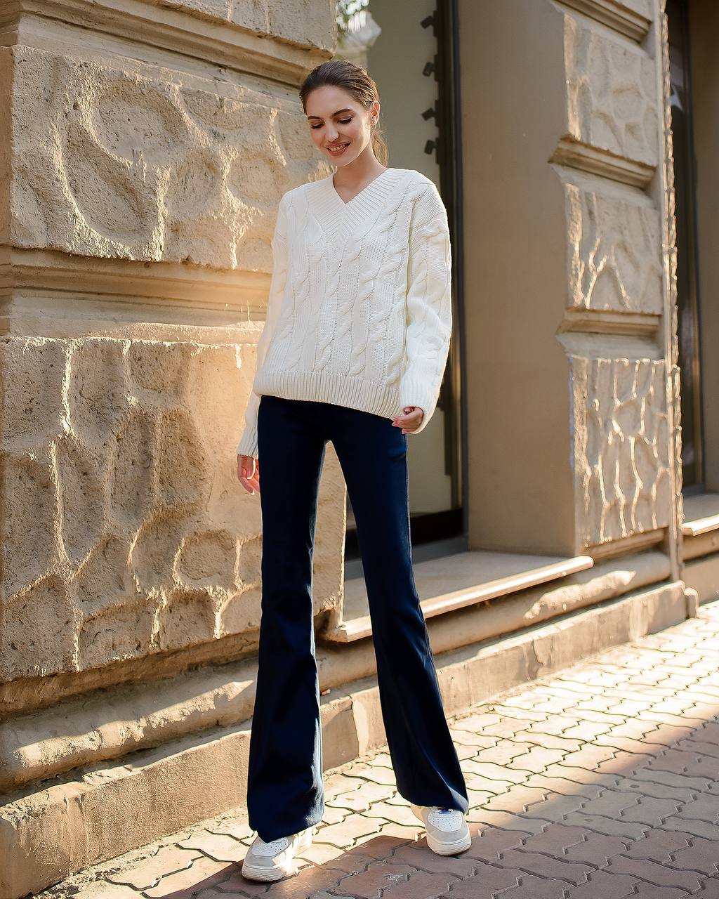 Woman wearing a white cable knit sweater and dark blue jeans standing in front of a stone building.