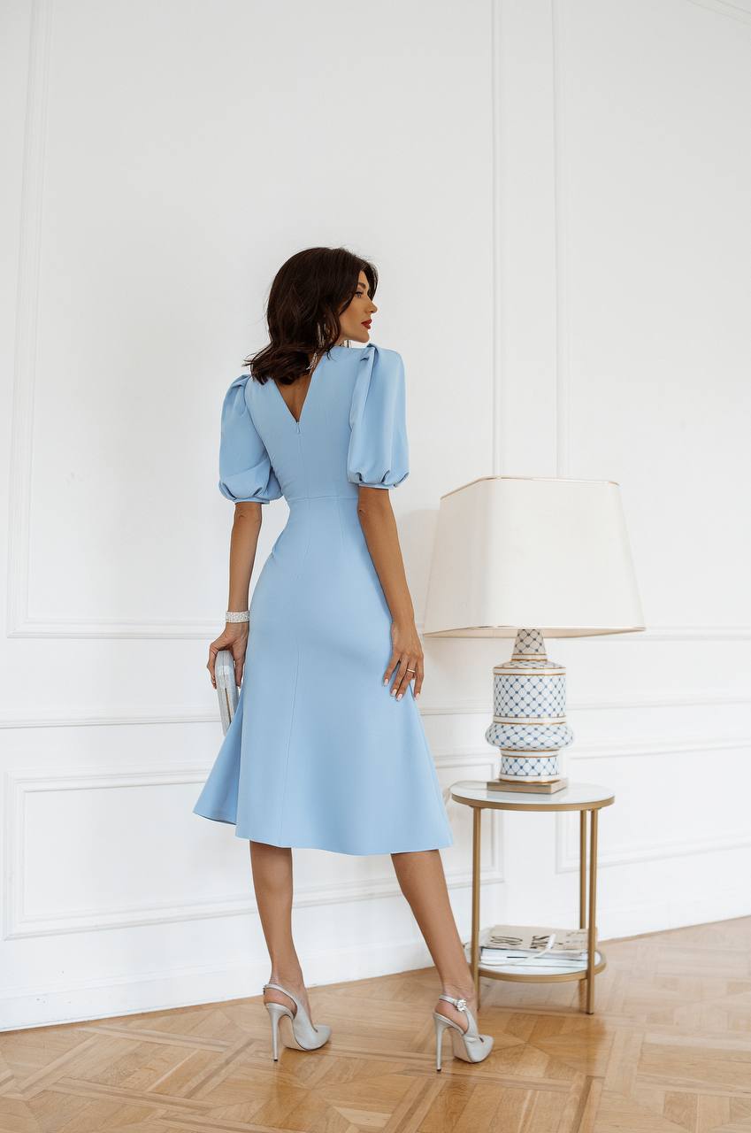Woman in a light blue dress standing in a room with a lamp and side table.
