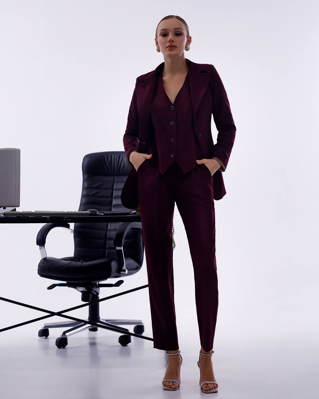 Woman in a burgundy suit standing in an office setting with a chair and desk.