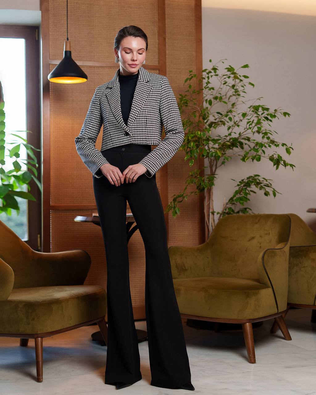 Woman in a checkered blazer and black pants standing in a modern living room.