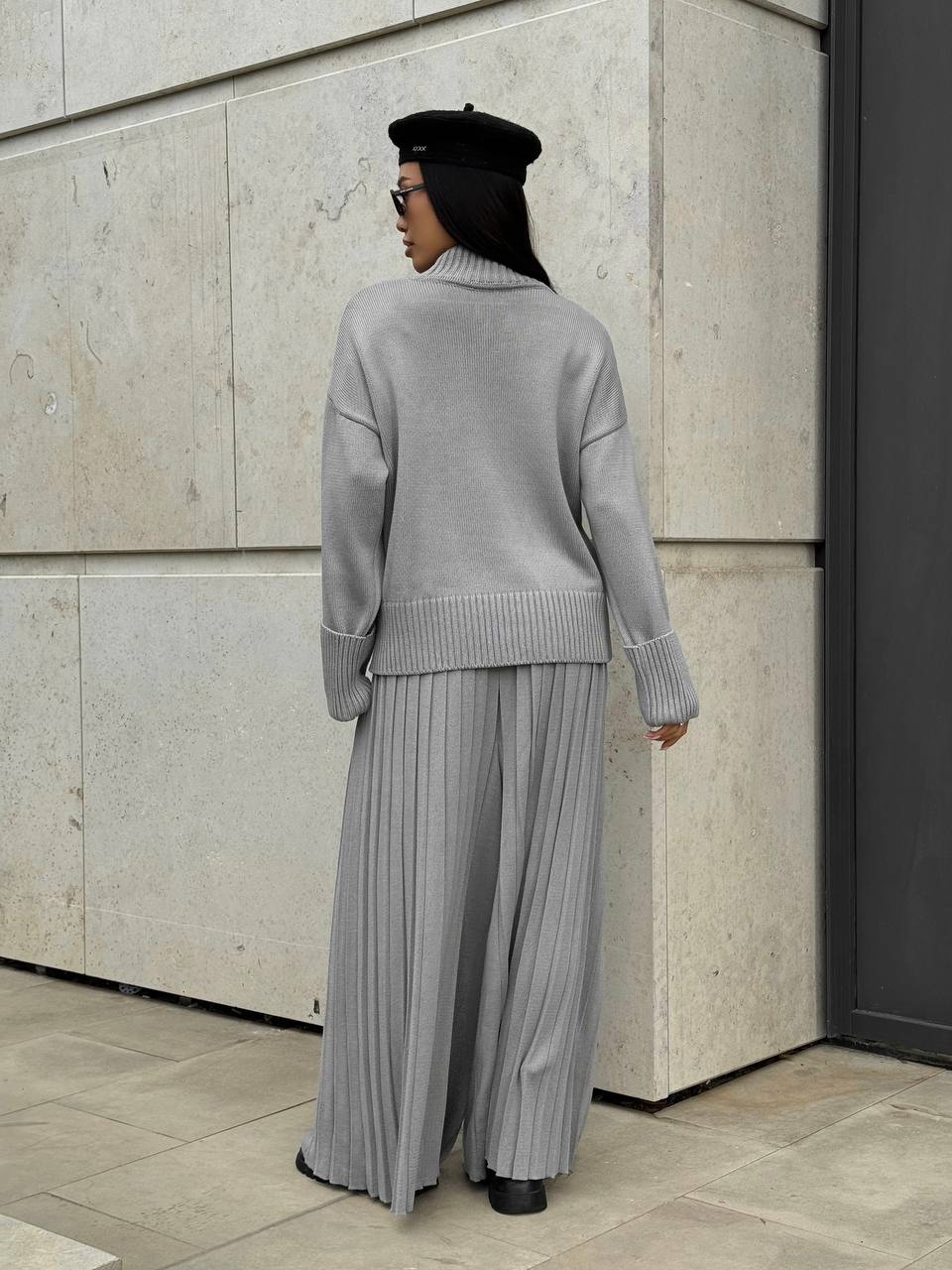 Person wearing a gray outfit with a pleated skirt against a concrete wall.