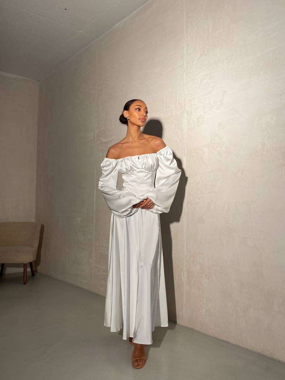 Woman in a white off-shoulder dress standing against a plain wall.