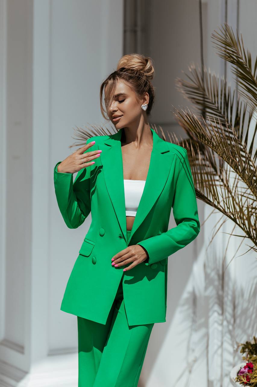 Woman wearing a green suit with a white top, standing against a light-colored wall with decorative elements.