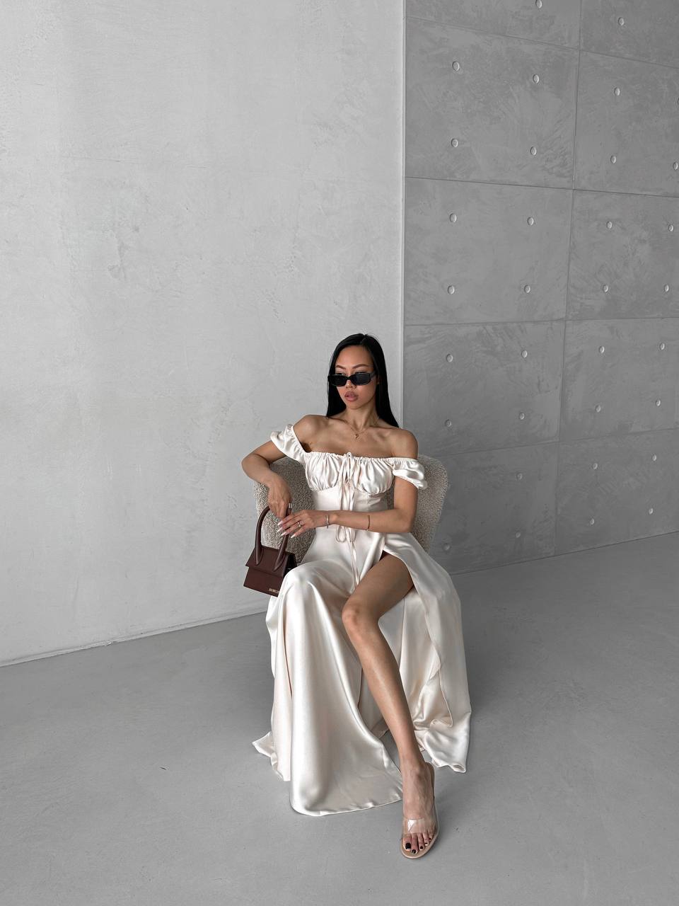 Woman in a white off-shoulder dress sitting on a chair against a gray wall.