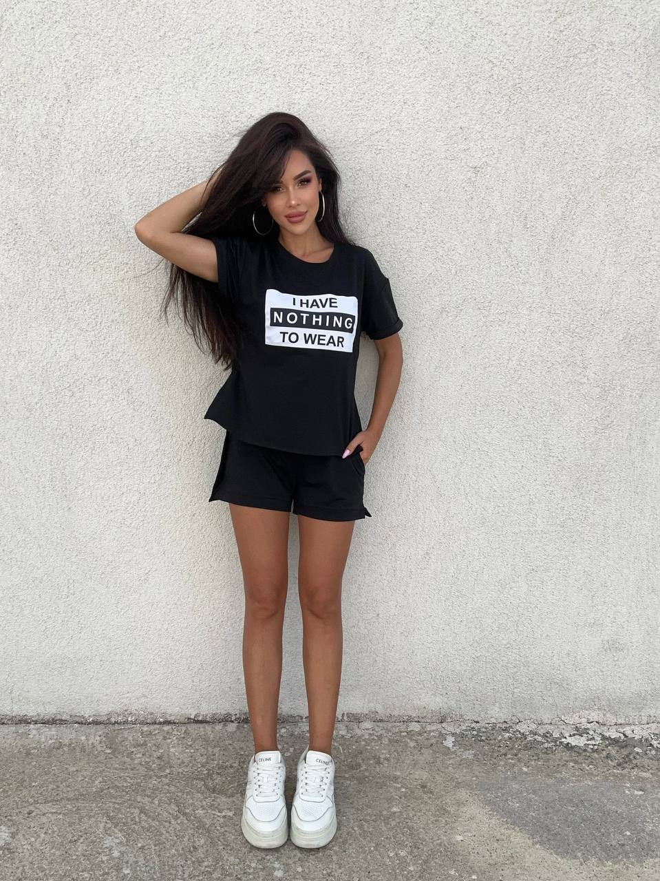 Person wearing a black t-shirt with text and shorts, standing against a plain wall.