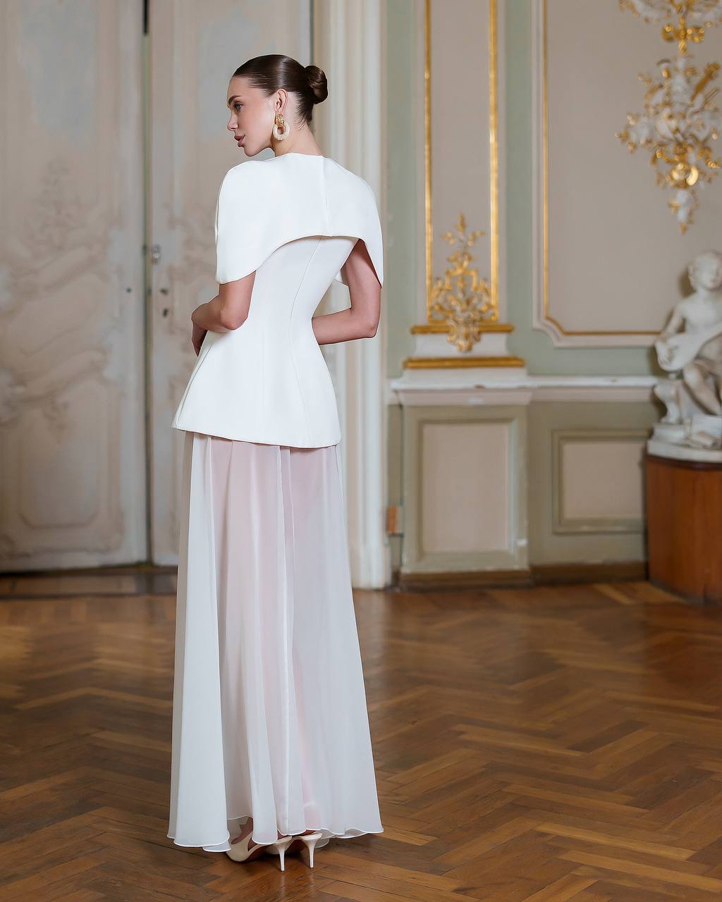 Woman in a white outfit standing in an elegant room with wooden floor and decorative wall.