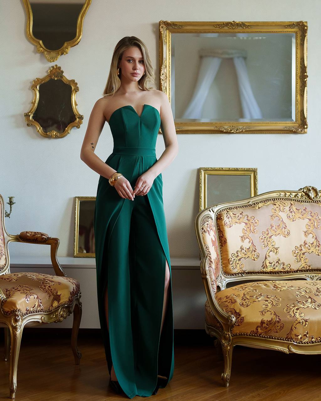 Woman in a green strapless gown standing in an elegant room with gold-framed mirrors and a decorative chair.