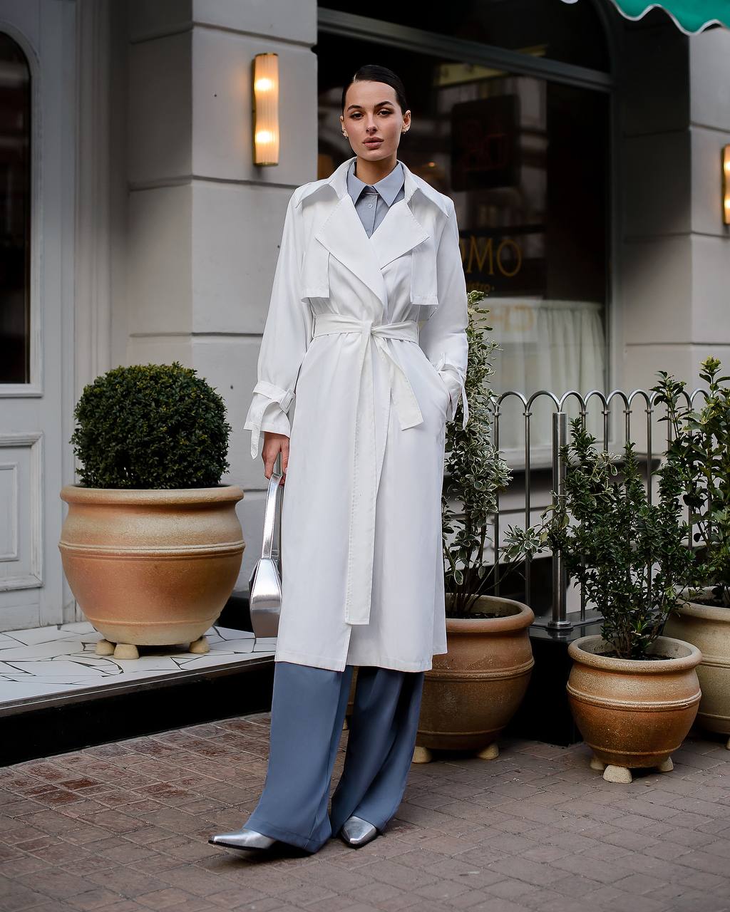 Woman in a white trench coat and blue pants standing in front of a building with plants.