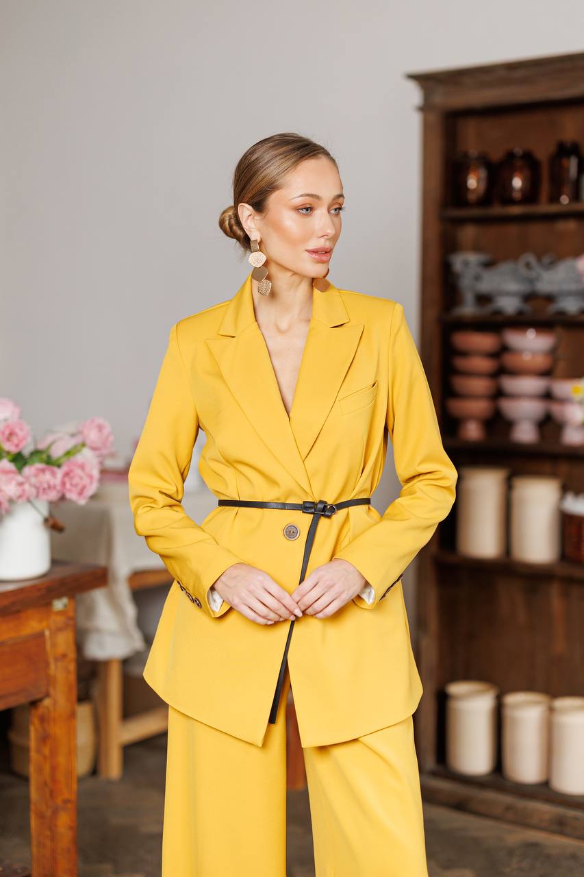 Woman in a yellow suit standing in a room with wooden furniture and decorative items.