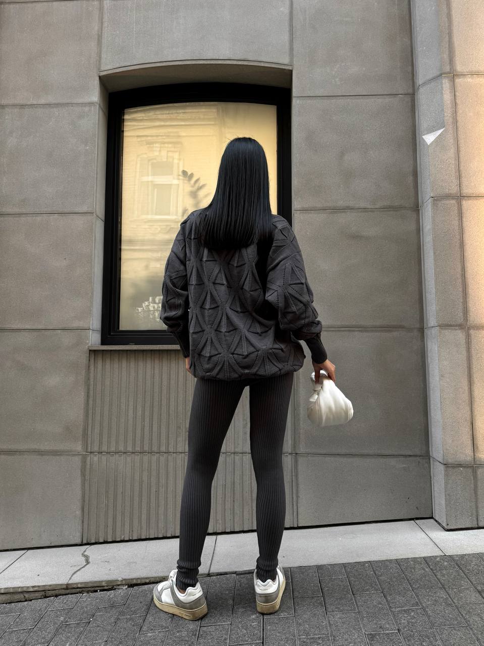 Person wearing a black quilted jacket and dark pants standing in front of a building with a window.