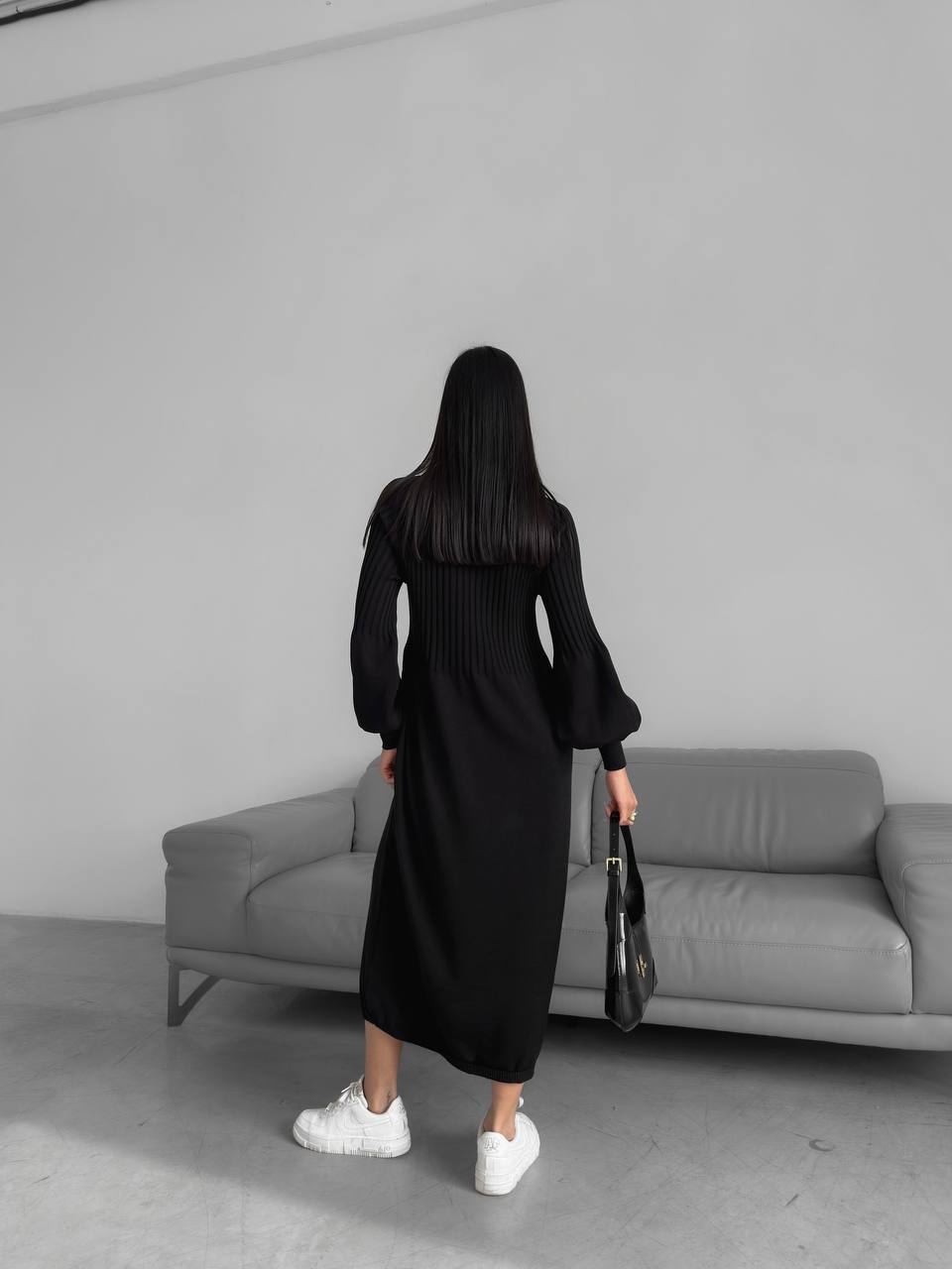 Person wearing a black dress standing in front of a gray sofa against a plain wall.