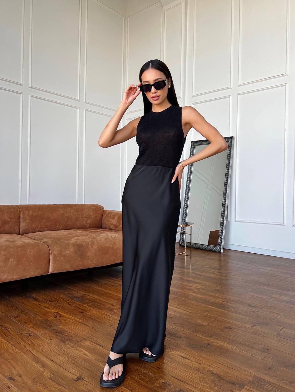 Woman in a black sleeveless top and wide-leg pants standing in a room with wooden flooring and a brown sofa.