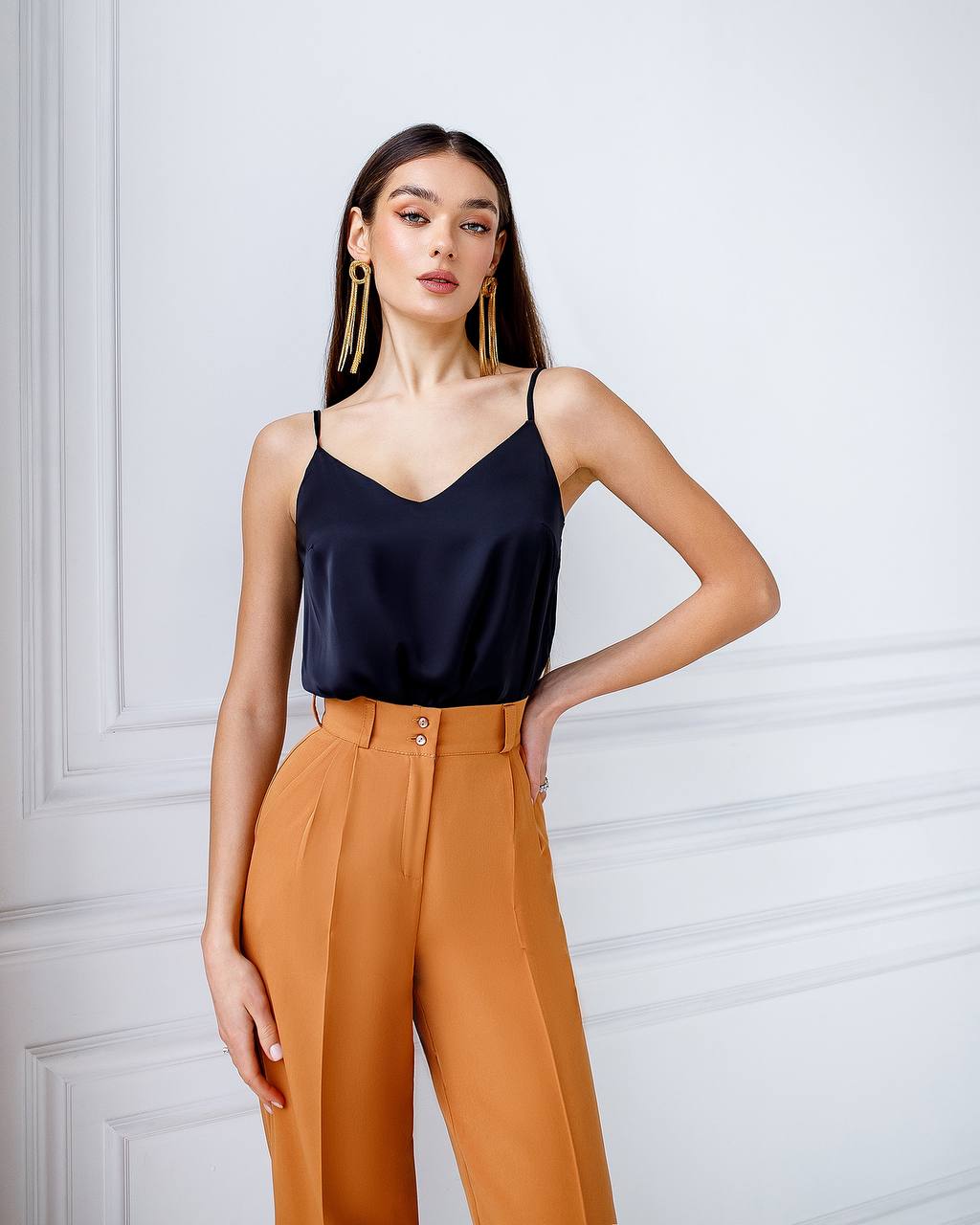 Woman wearing a black top and orange pants against a white wall