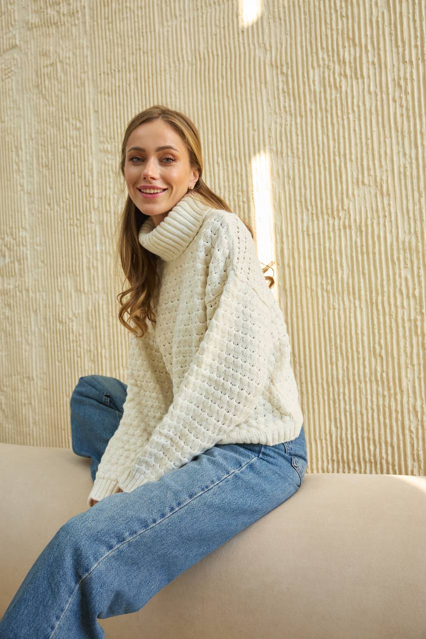 Woman wearing a white sweater and blue jeans sitting on a couch against a textured beige wall.
